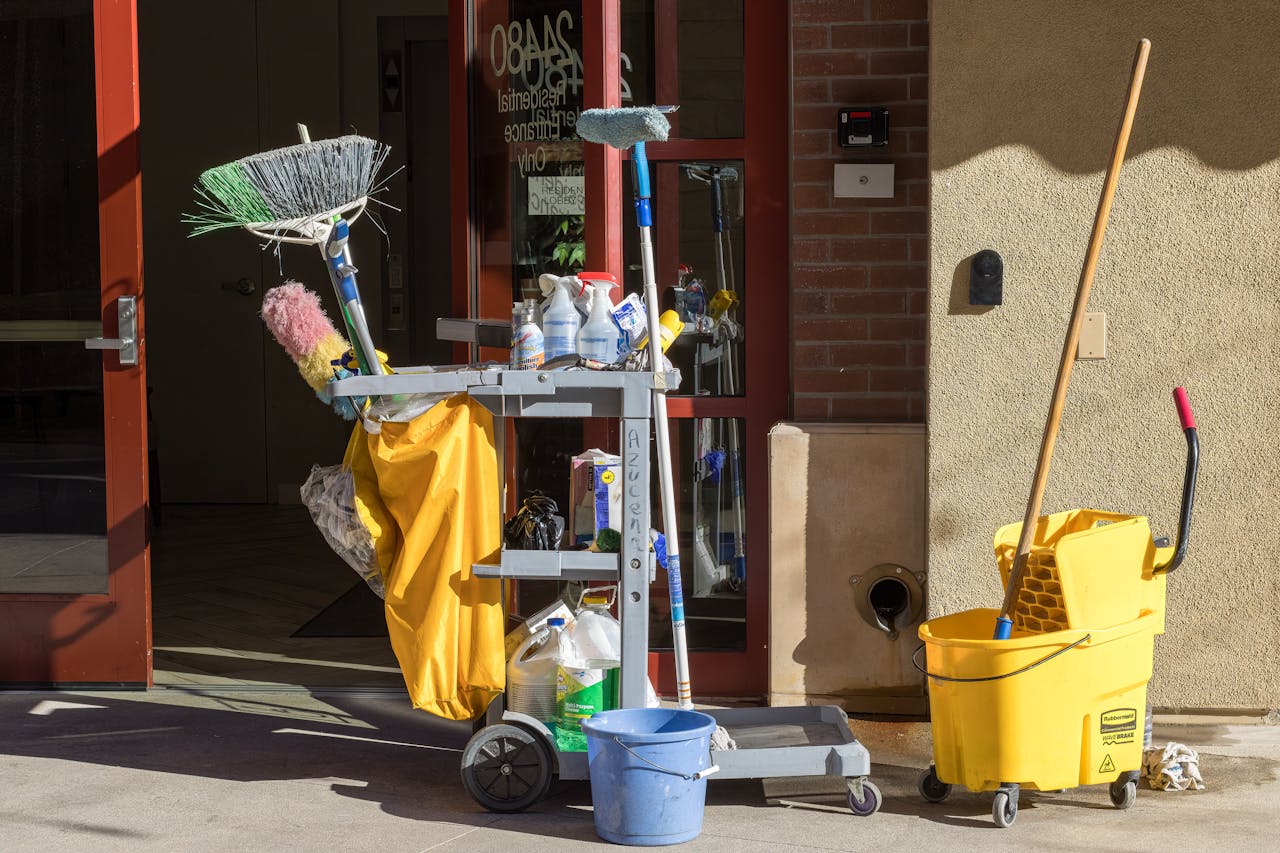 gallery-6 Cleaning supplies and tools outside a building entrance bathed in sunlight.