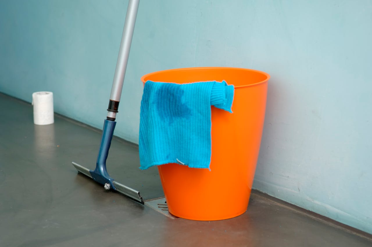 hero-img-01 Close-up of cleaning supplies including orange bucket, mop, and blue rag indoors.